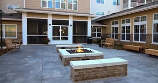 Outdoor courtyard area with a central fire pit surrounded by brick benches and wooden benches along the building walls. The courtyard is enclosed by a multi-story building with large windows and screened doors.