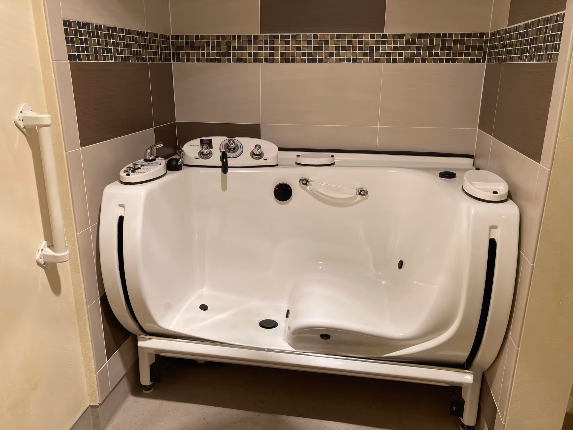 A white walk-in bathtub with built-in seat and multiple control knobs, installed in a tiled bathroom corner with beige and brown tiles. There is a white grab bar mounted on the left wall.