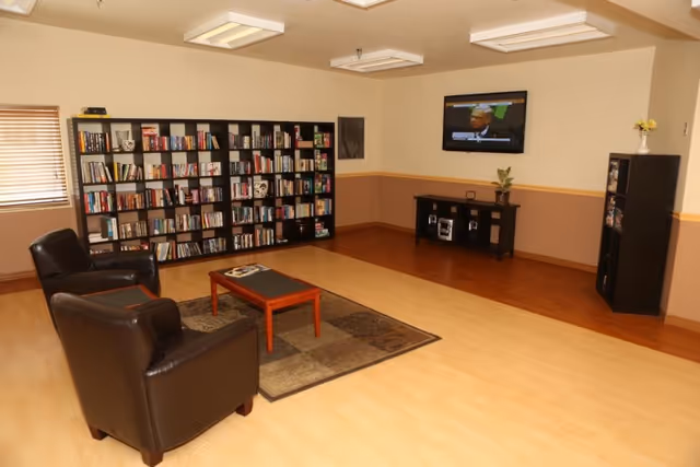 A cozy living room area with two black leather armchairs facing a small wooden coffee table on a patterned rug. Behind the chairs is a large bookshelf filled with books. A flat-screen TV is mounted on the wall above a small black entertainment console with decorative items. The room has light-colored walls, wooden flooring, and a window with blinds on the left side.
