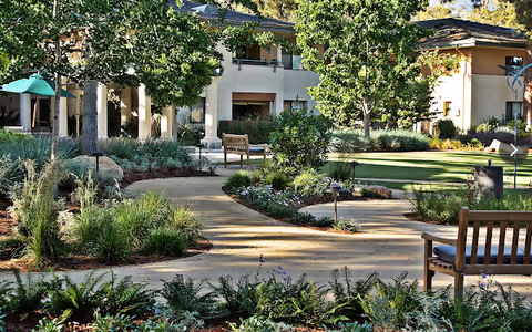 A landscaped outdoor garden area at Vista del Monte Retirement Community featuring winding pathways, green lawns, various plants and trees, wooden benches, and a building with balconies in the background.