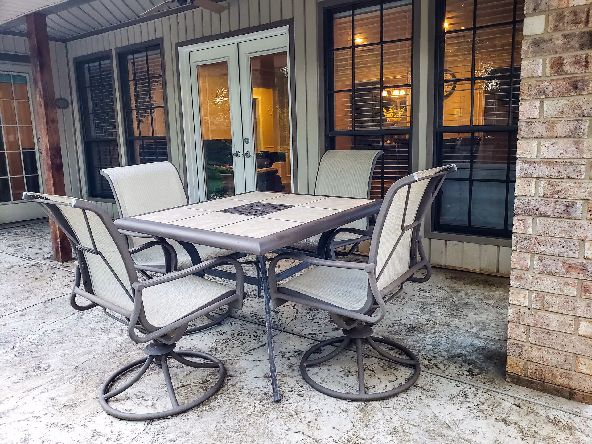 Outdoor covered patio with a square tile-top dining table and metal-framed swivel chairs in front of glass doors and windows.