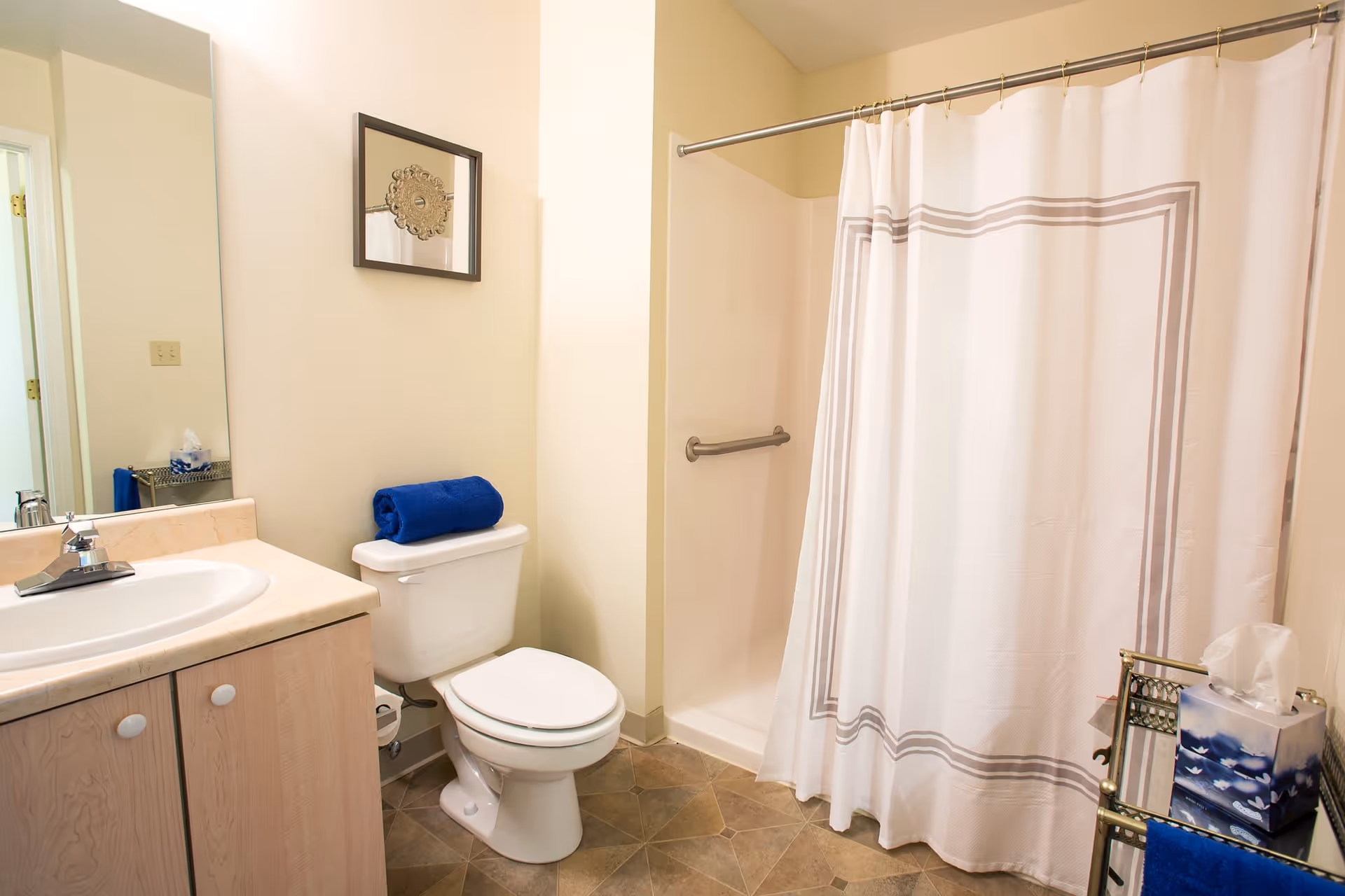 A clean bathroom featuring a white toilet with a blue towel rolled on top, a sink with a beige countertop and wooden cabinet, a shower with a white curtain and a grab bar, a tissue box on a small metal rack, and a framed decorative mirror on the wall.