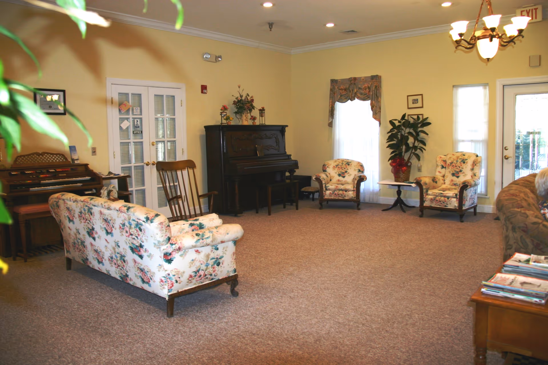 A spacious living room with floral upholstered sofa and armchairs, a wooden rocking chair, a piano, an organ, a small table with a plant, and a chandelier. The walls are painted yellow, and there are windows with curtains and a glass door leading outside.