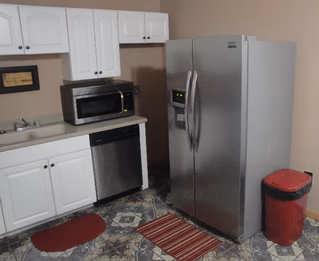 A kitchen area with white cabinets, a stainless steel microwave, dishwasher, and a large stainless steel refrigerator. The floor has patterned tiles with two small rugs, one red and one striped. There is a red trash bin in the corner.
