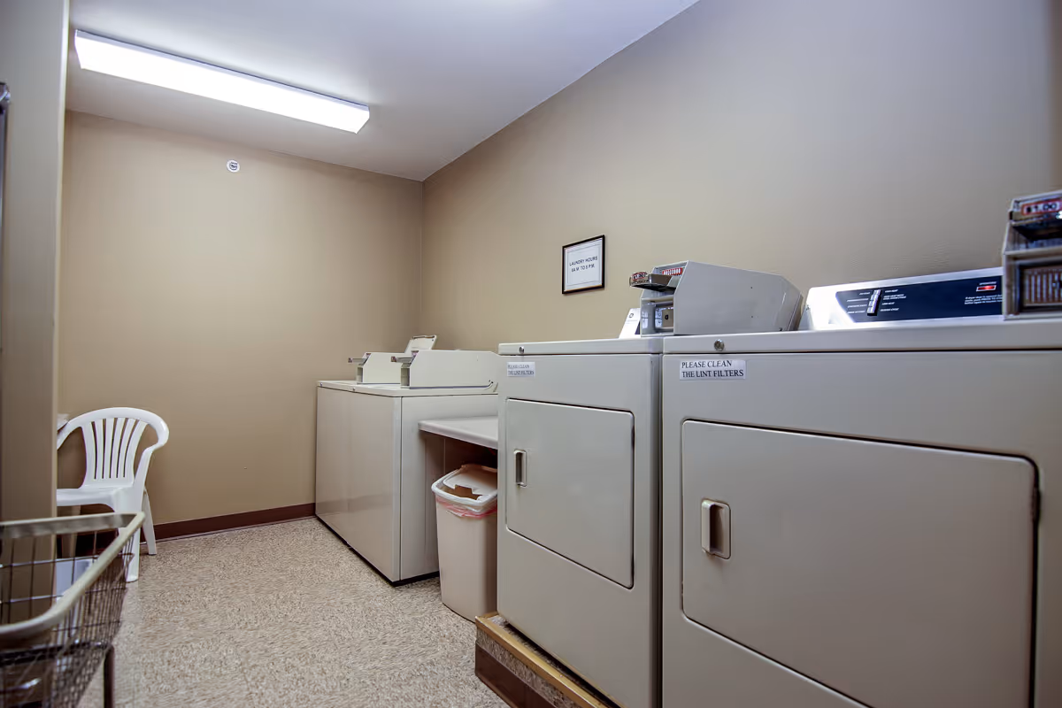 Laundry room with beige walls and a fluorescent ceiling light. The room contains two washing machines and two dryers, a white plastic chair in the corner, a trash can, and a laundry cart. There are small signs on the machines and a framed sign on the wall.
