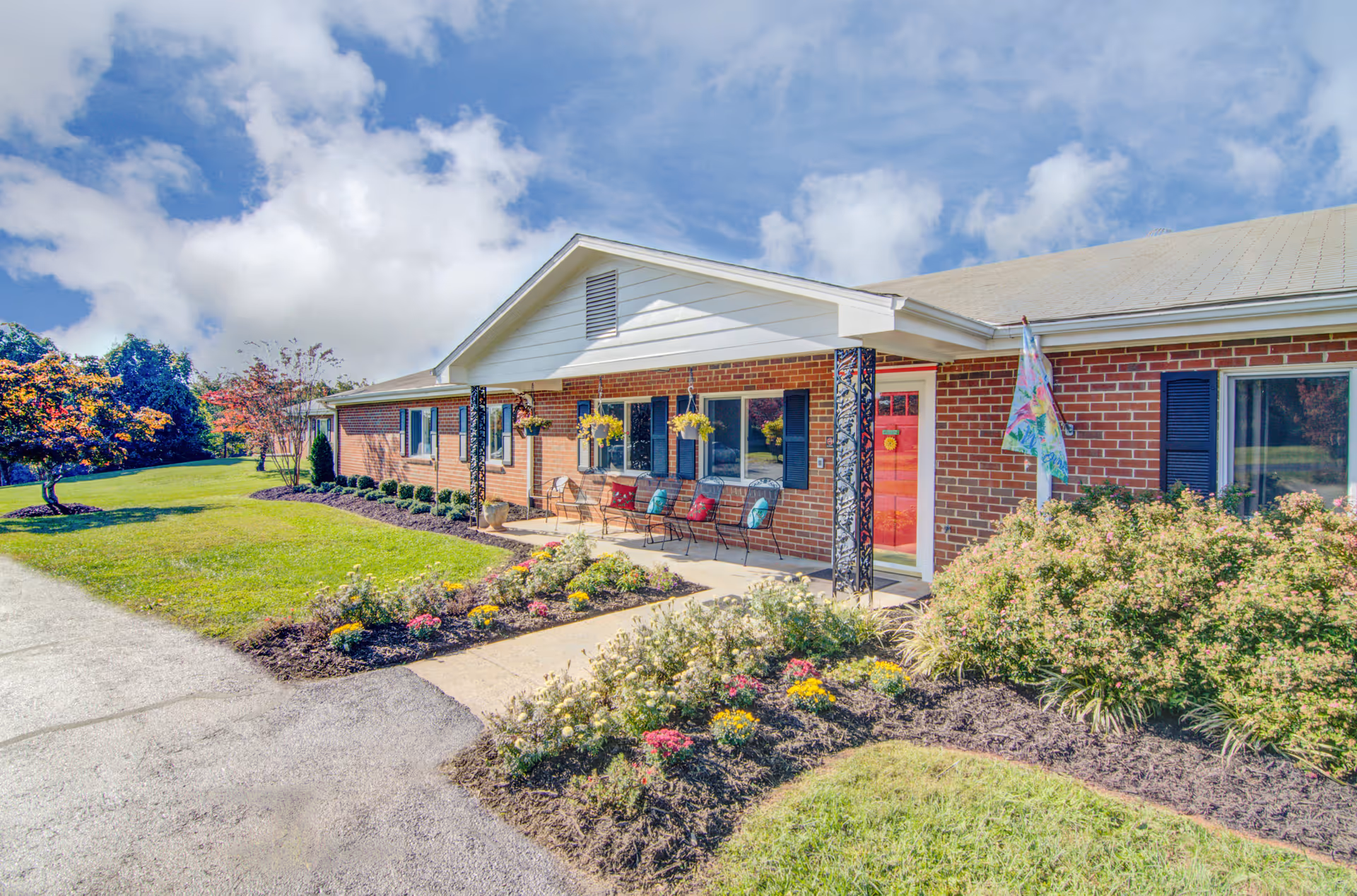 Exterior view of a single-story brick building with a white roof and a red front door. The building has black shutters on the windows and a small covered porch with decorative black metal posts and several chairs with colorful cushions. The front yard is landscaped with green grass, flowering plants, and shrubs under a partly cloudy blue sky.