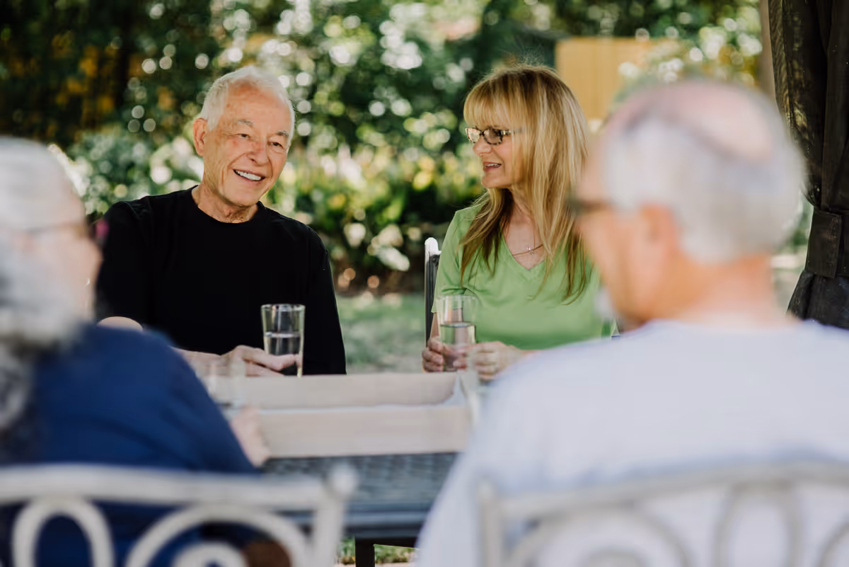 Four elderly people sitting outdoors around a table, two of them holding glasses of water, engaged in conversation with greenery in the background.