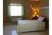 A simple bedroom with a single bed covered in a patterned bedspread, a pillow, a wooden nightstand with a lamp, a colorful wall decoration, a window with green curtains, and a heating or air conditioning unit below the window.