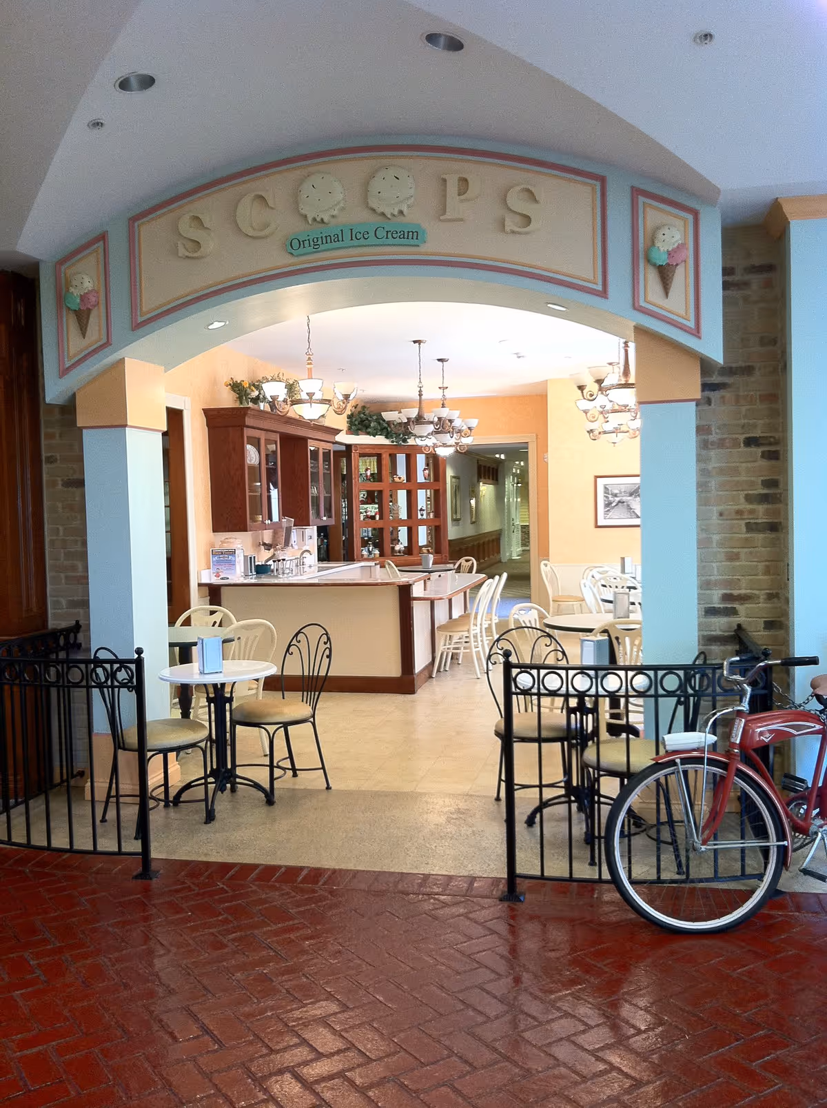 Interior view of an ice cream parlor named 'Scoops Original Ice Cream' with tables and chairs arranged for seating. The area has a counter with cabinets and chandeliers hanging from the ceiling. A red bicycle is parked near the entrance gate.