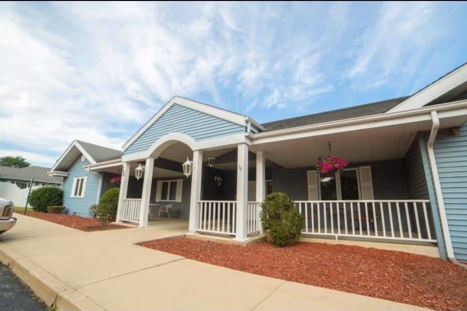 Exterior view of a single-story building with blue siding and white trim, featuring a covered porch with white railings and hanging lanterns. There are bushes and a flower pot with pink flowers near the porch, and a concrete walkway in front.