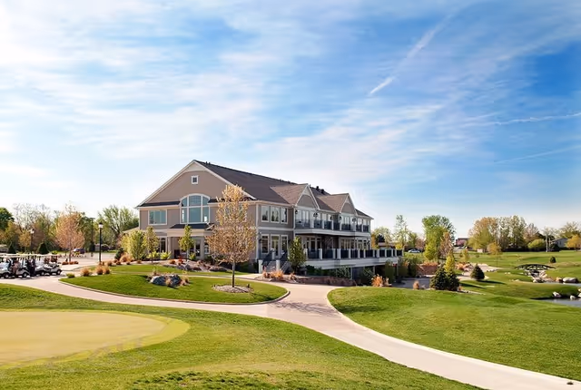 A large two-story building with a beige exterior and multiple windows, surrounded by well-maintained green lawns, trees, and a paved walkway under a partly cloudy blue sky.