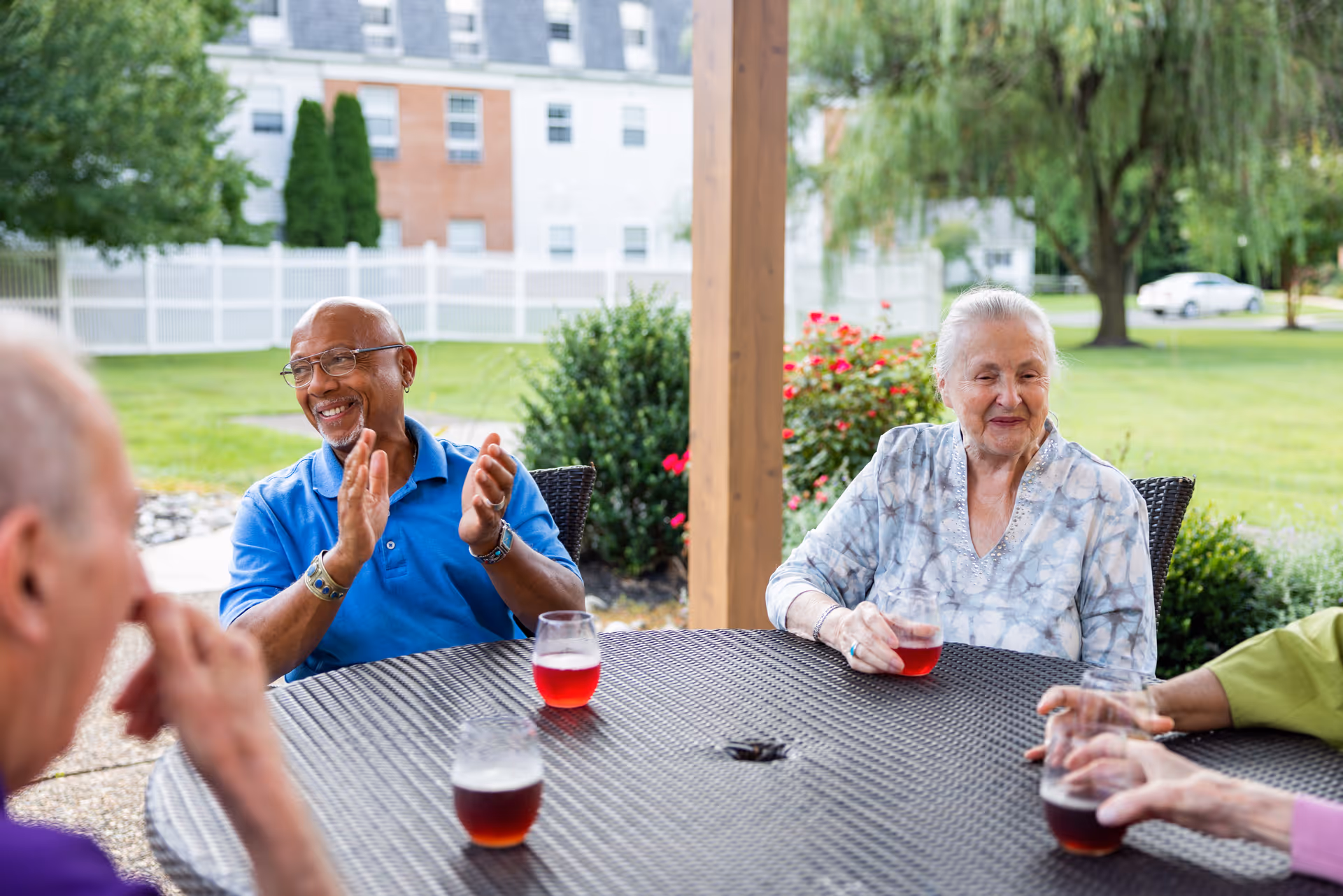 A group of elderly people sitting around a round outdoor table with drinks, enjoying a sunny day in a garden area with greenery and flowers in the background.