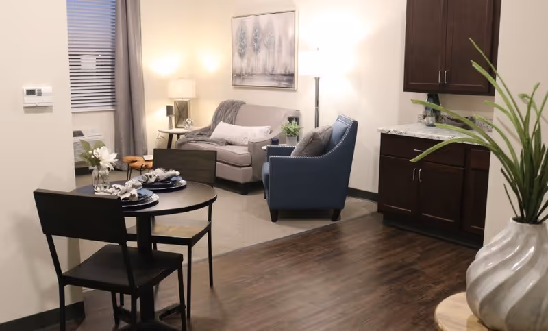 A cozy living area with a small round dining table set for two, a gray sofa with a throw blanket and pillow, a blue armchair, a side table with a lamp, a piece of wall art depicting trees, and a kitchenette with dark wood cabinets and a granite countertop. A potted plant is visible in the foreground.
