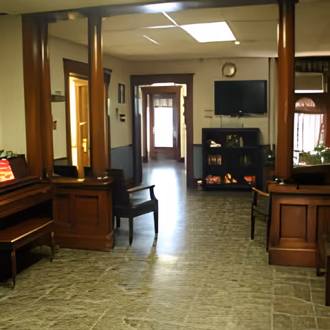 Interior view of a senior living facility common area with wooden pillars, chairs, a piano with a red lamp, a TV mounted on the wall, and a bookshelf. The floor has a stone-like pattern and there is natural light coming from a doorway in the background.
