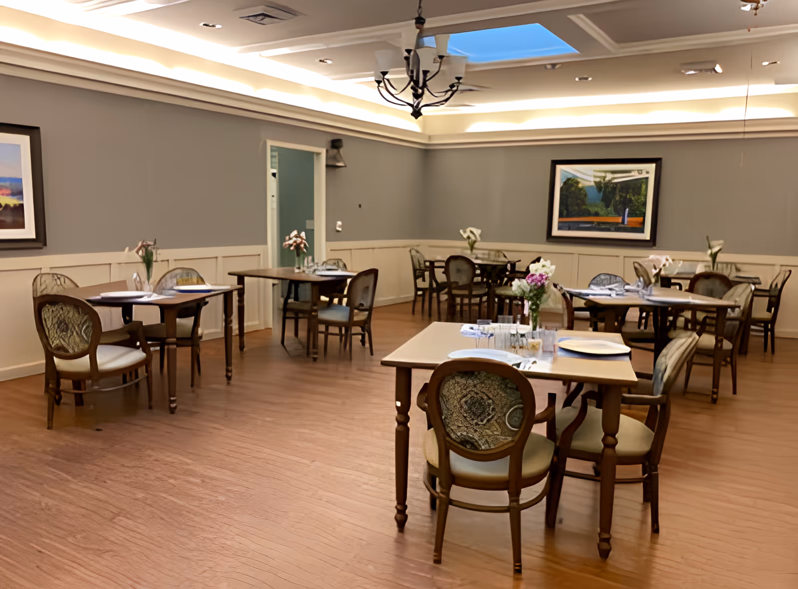 A dining room with several wooden tables and upholstered chairs arranged neatly. Each table has a vase with flowers and place settings including plates, glasses, and napkins. The room has wooden flooring, gray walls with white wainscoting, framed artwork on the walls, and a ceiling light fixture with a skylight.