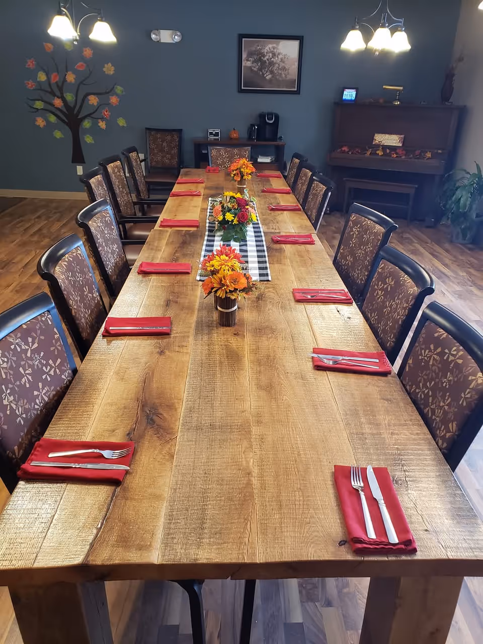 Long wooden dining table set for a meal with chairs, red napkins and floral centerpieces in a communal dining room.