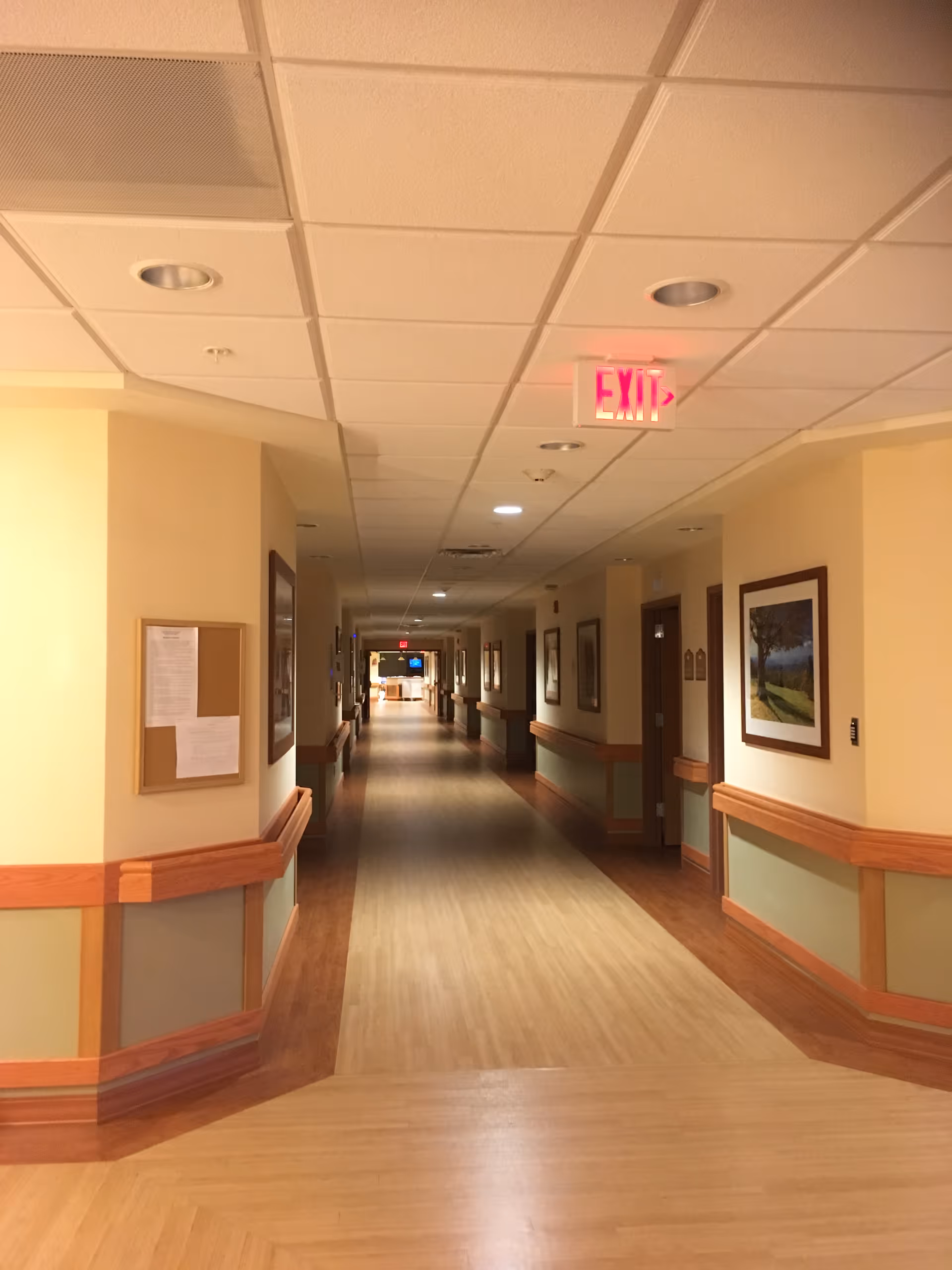 A long, well-lit hallway in a senior living facility with light-colored walls, wooden handrails, framed pictures on the walls, and an exit sign on the ceiling.