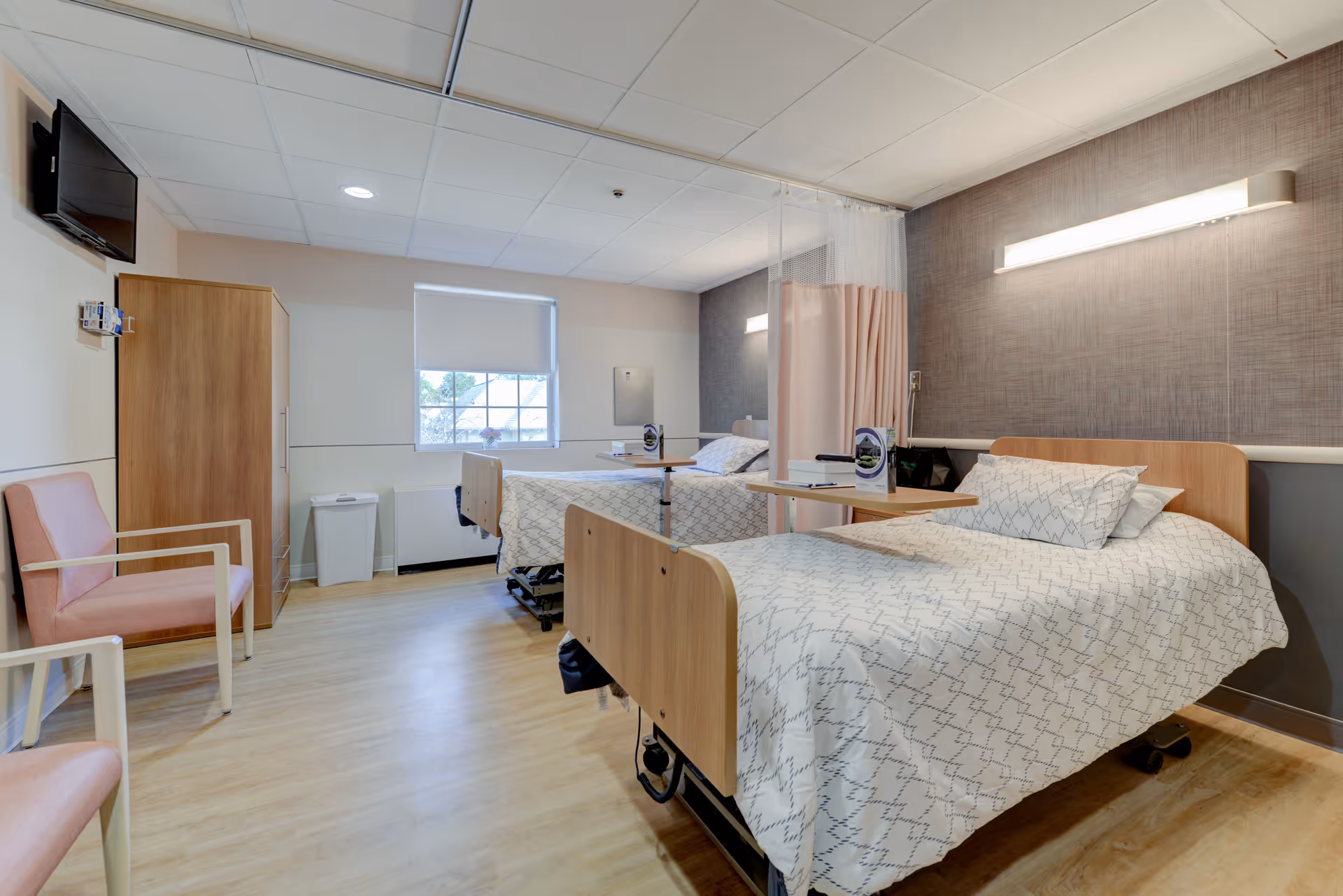 A clean and well-lit hospital or nursing facility room with two beds separated by a privacy curtain. Each bed has white patterned bedding and an overbed table with brochures. The room features light wood flooring, a wooden wardrobe, two pink chairs, a wall-mounted TV, and a window with a roller shade partially down.