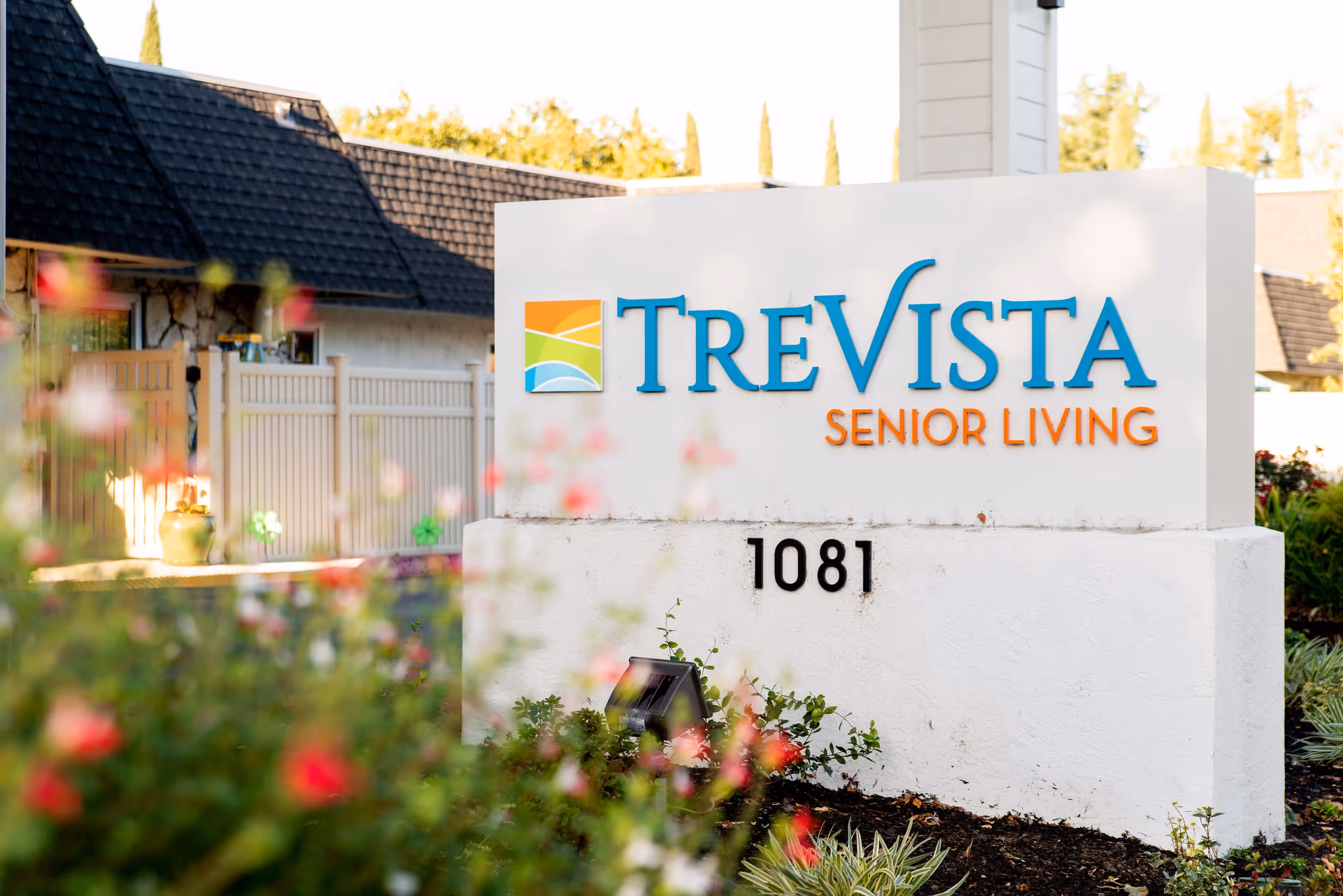 Outdoor view of a white sign with the colorful TreVista logo and the text 'TreVista Senior Living' along with the number 1081, surrounded by greenery and flowers with a building and fence in the background.