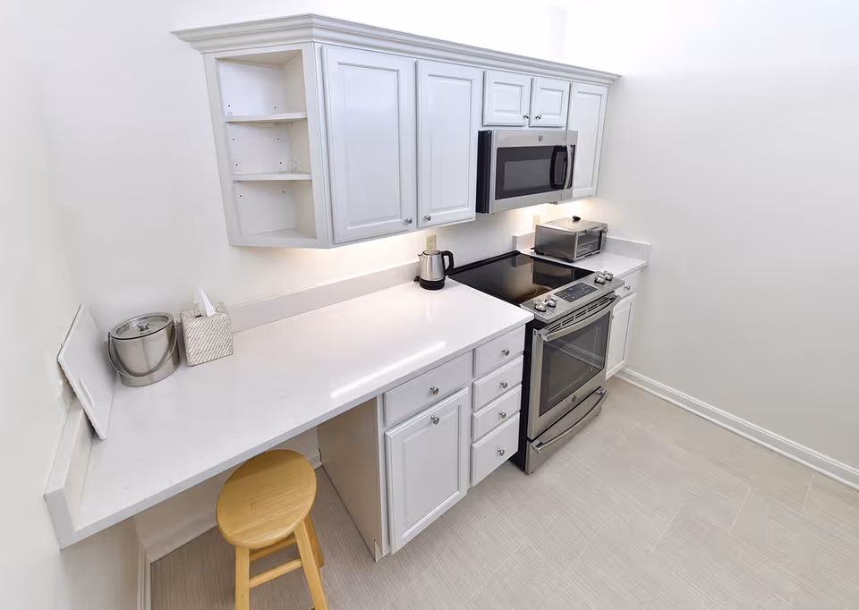 A clean, modern kitchen with white cabinets and countertops. The kitchen features a stainless steel electric stove with an oven, a microwave mounted above the stove, a toaster oven, an electric kettle, and a tissue box on the counter. There is a wooden stool tucked under the counter, and the floor is covered with light-colored tiles.
