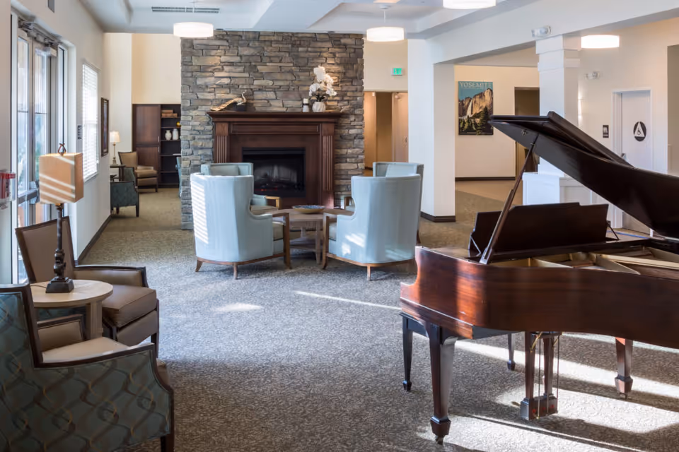 A spacious and well-lit common area in a senior living facility featuring a grand piano on the right, a stone fireplace with a wooden mantel in the center, and several comfortable chairs arranged around small tables. The room has carpeted floors, large windows on the left letting in natural light, and neutral-colored walls with some framed artwork.