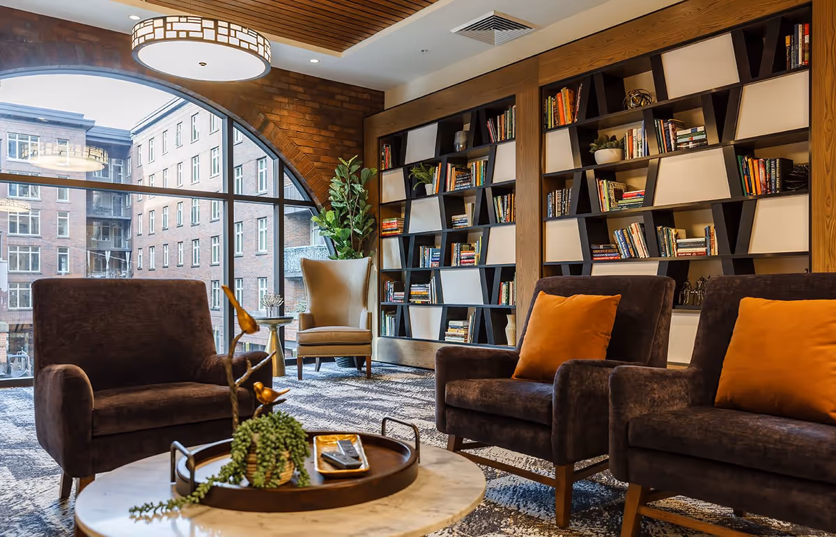 A cozy living room area with large arched windows showing a view of brick buildings outside. The room features a modern bookshelf filled with books, two dark brown armchairs with orange cushions, a beige wingback chair, a round marble coffee table with a decorative tray and plant, and a large ceiling light fixture.