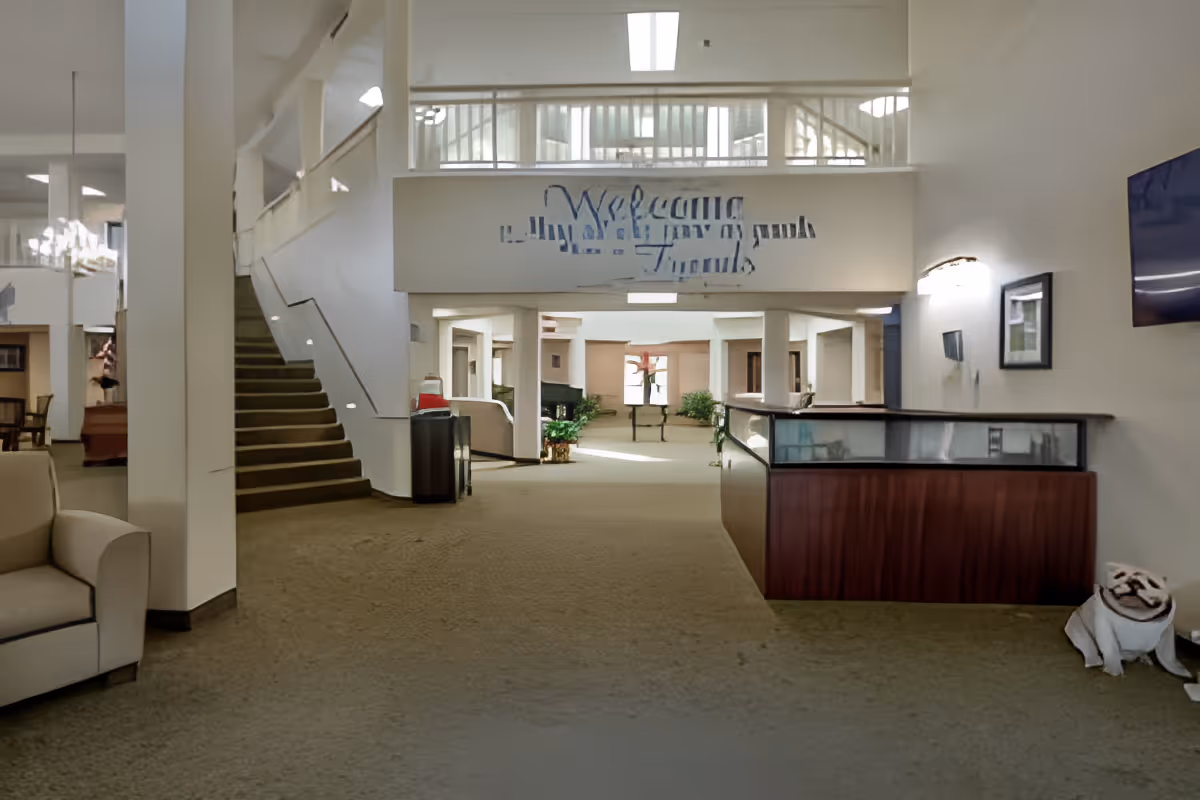 Spacious senior living facility lobby with a reception desk, staircase, seating, and a 'Welcome' sign on the far wall.