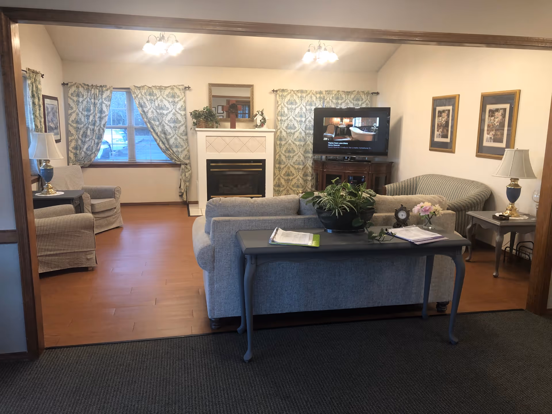 A cozy senior living facility common area with a gray sofa, two armchairs, a loveseat, and a wooden table with plants and paperwork. The room features a fireplace with a mirror above it, a TV on a wooden stand, floral curtains on the windows, and framed floral artwork on the walls. Two table lamps with white shades are placed on side tables beside the seating.