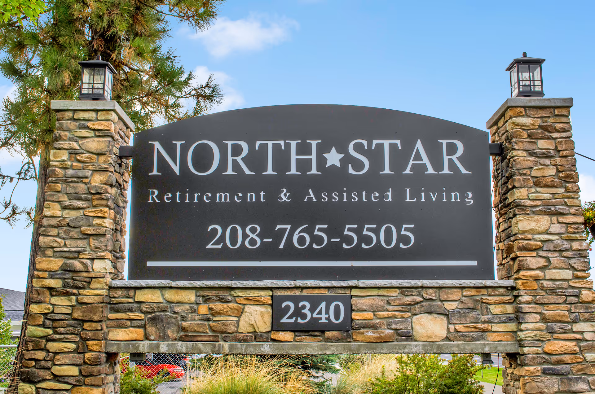 Stone signpost with lanterns on top displaying the name North Star Retirement & Assisted Living with a phone number 208-765-5505 and the address number 2340, set against a background of trees and blue sky.