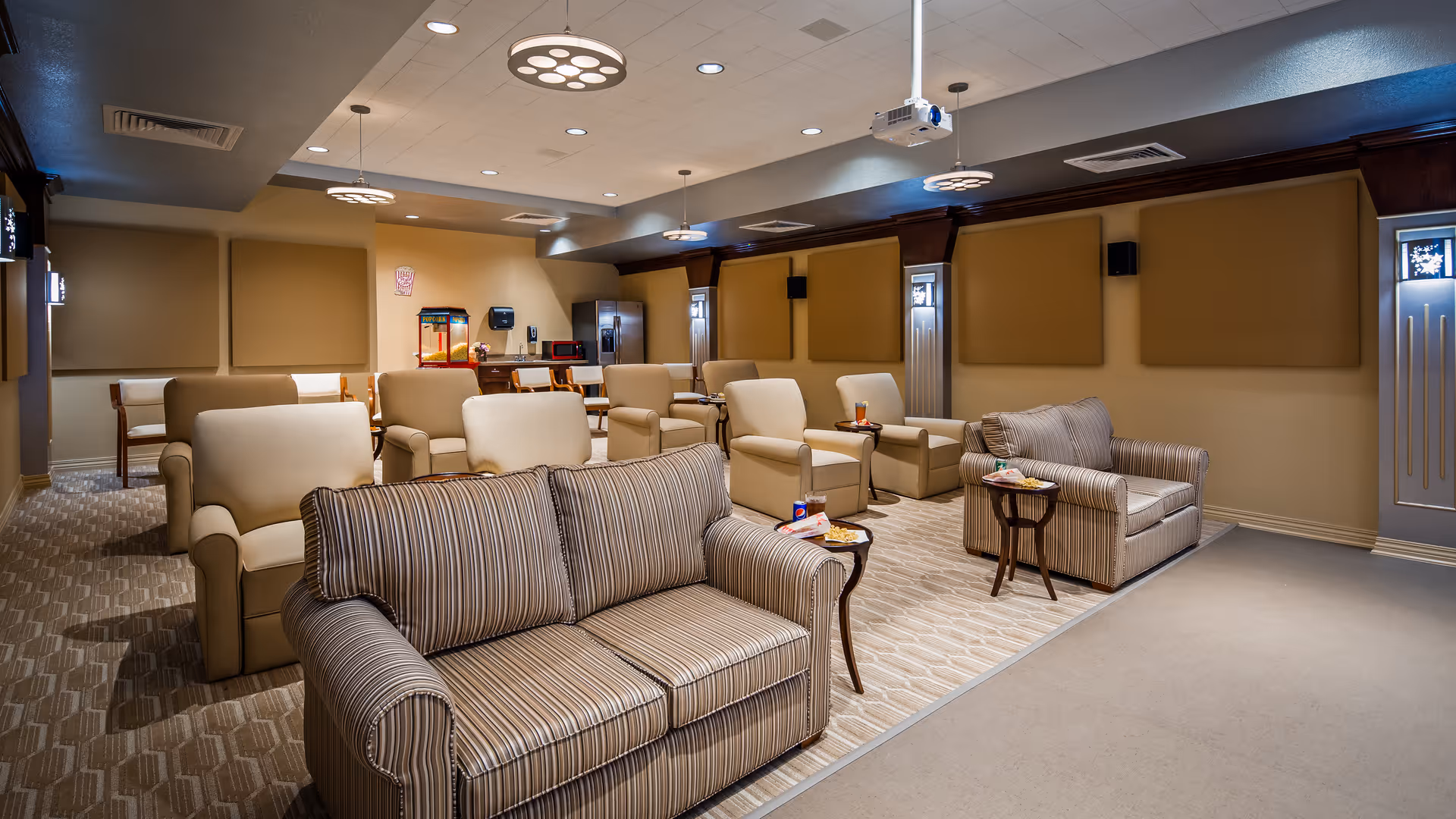 A cozy common area with multiple beige armchairs and striped sofas arranged in rows facing a blank wall, with small side tables holding snacks and drinks. The room has soft lighting from ceiling fixtures, beige walls with padded panels, and a carpeted floor. In the background, there is a kitchenette area with a refrigerator, microwave, and a popcorn machine.