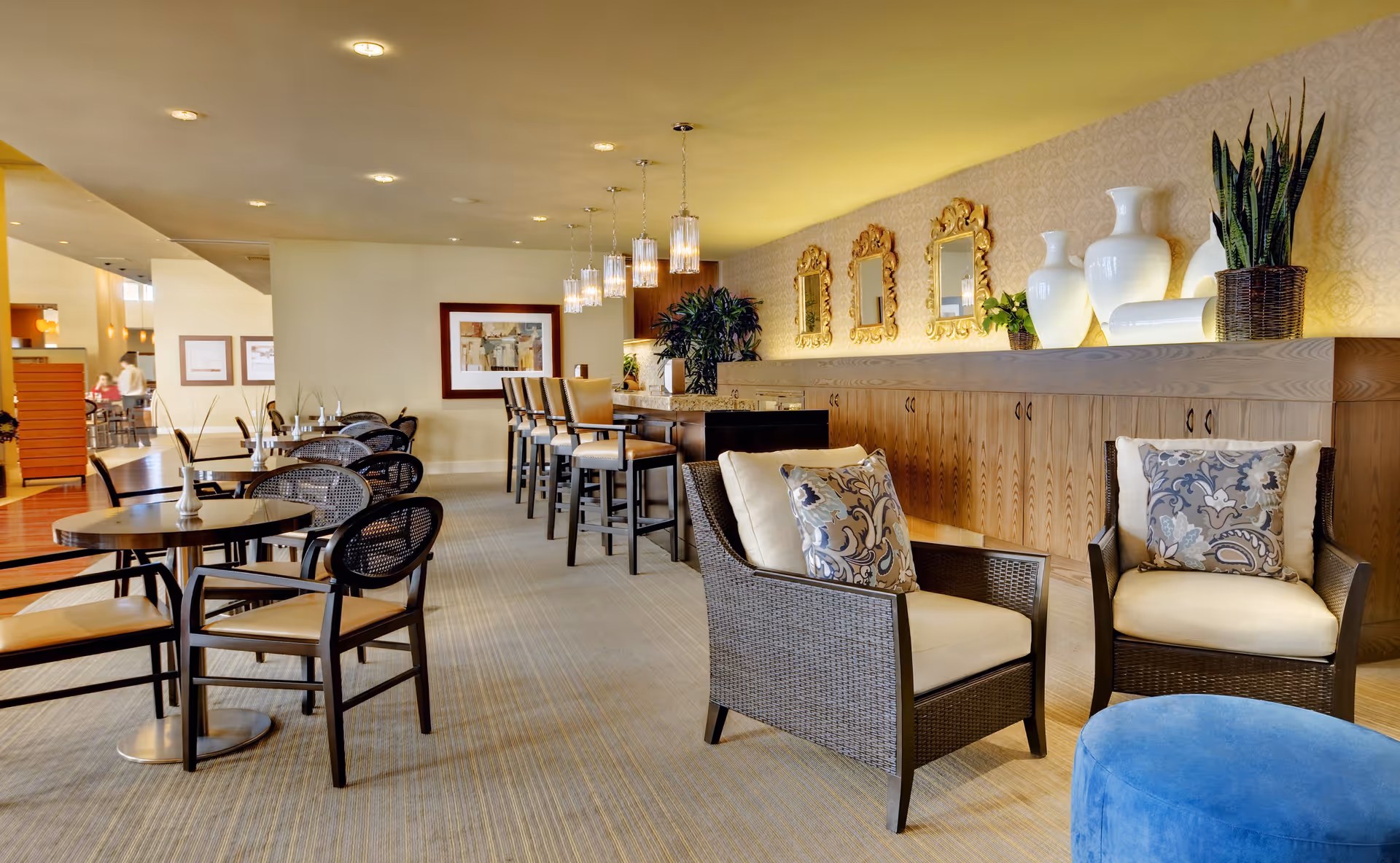 A spacious and well-lit common area with multiple seating options including cushioned armchairs with patterned pillows, round tables with chairs, and a high counter with bar stools. The wall behind the counter features decorative gold-framed mirrors, large white vases, and potted plants. The room has a warm and inviting ambiance with soft lighting and carpeted flooring.