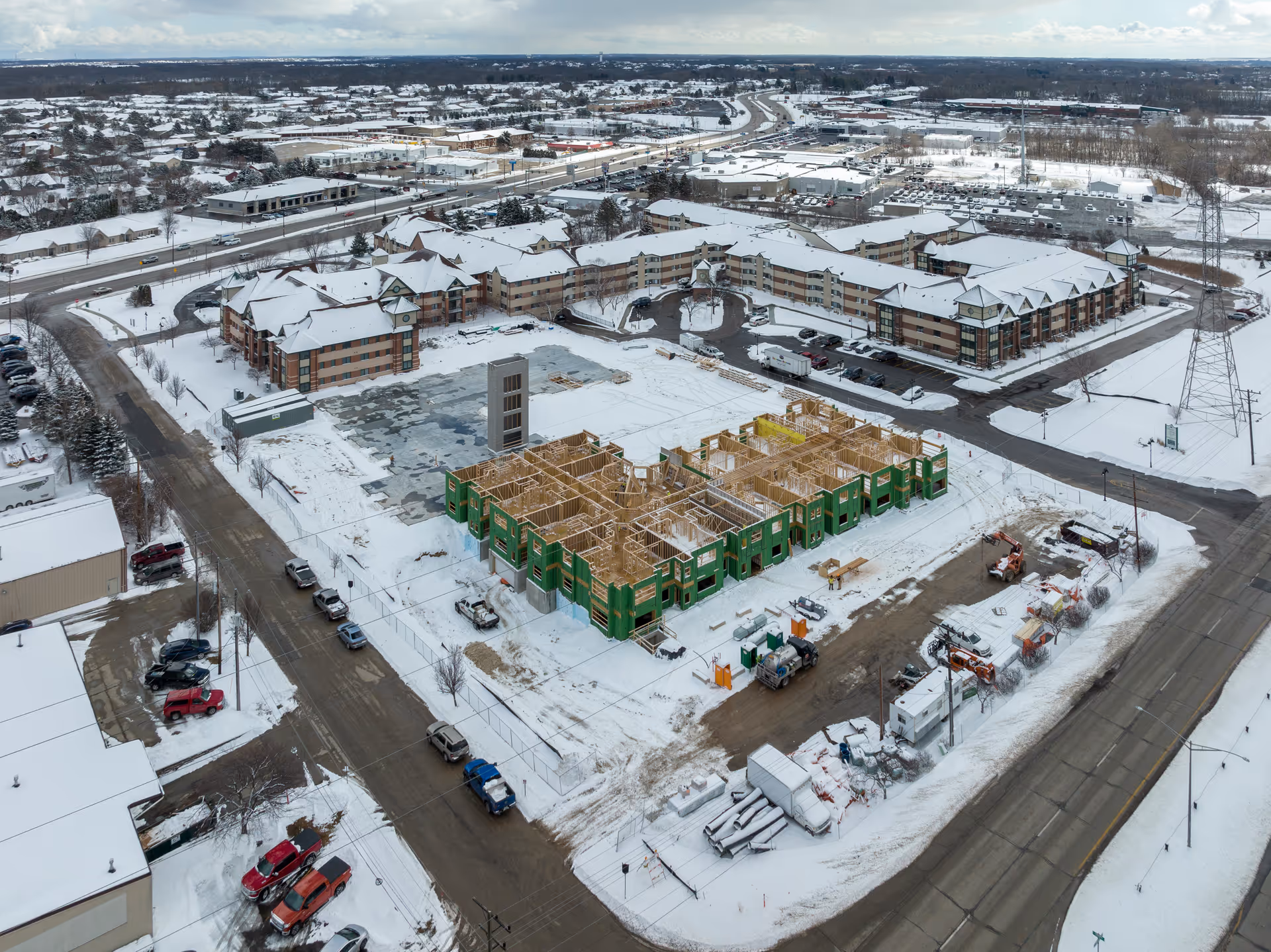 Aerial view of a senior living community named Forest Ridge Senior Community during winter. The image shows multiple large buildings with snow-covered roofs surrounding a construction site where a new building is being framed. Snow covers the ground and roads, with several vehicles parked along the streets and construction equipment visible near the site.