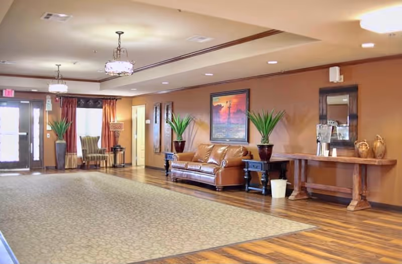 A spacious and well-lit common area with wooden flooring and a large patterned rug. The room features a brown leather sofa flanked by two potted plants on dark wooden side tables. A long wooden table against the wall holds a water dispenser and decorative items. The walls are painted a warm brown color and adorned with framed artwork and a mirror. Two chandeliers hang from the ceiling, and there are chairs near the entrance door with windows covered by curtains.
