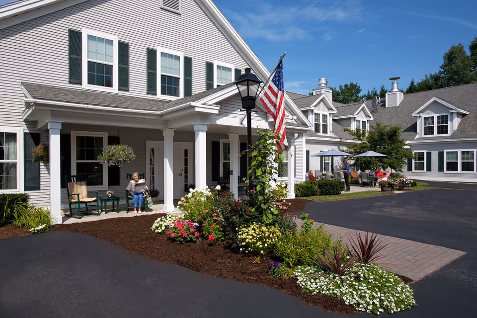 Exterior view of Woodstock Terrace senior living facility showing a large white building with green shutters, a covered porch with seating, an American flag on a lamp post, and landscaped flower beds. Several people are sitting and standing outside under umbrellas on a sunny day.