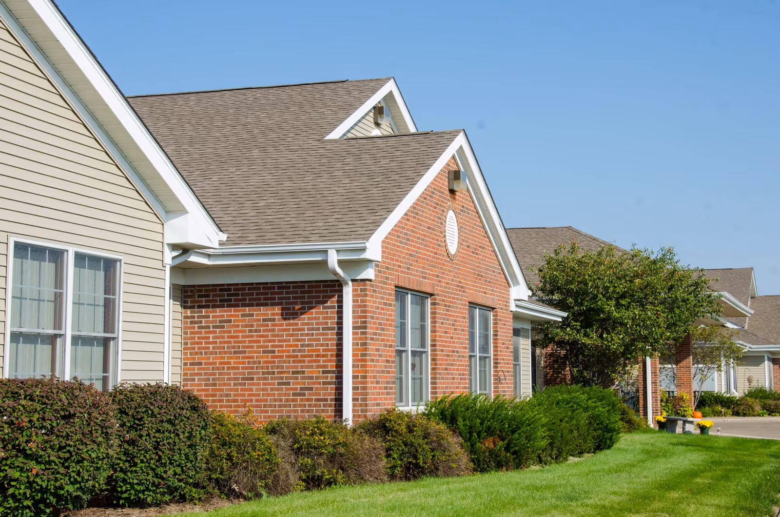 Exterior view of a single-story building with brick and beige siding, multiple windows, a well-maintained lawn, shrubs, and a tree under a clear blue sky.