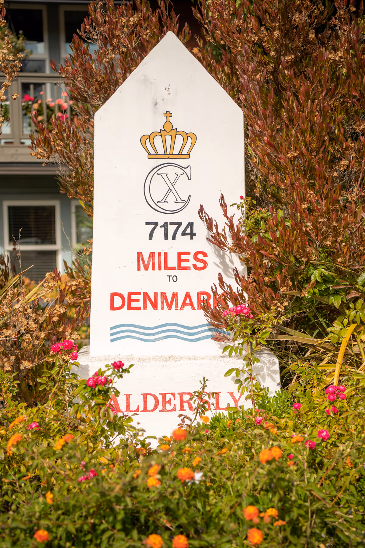 A white milepost-style sign in a flowering garden that reads '7174 MILES TO DENMARK' with a crown emblem and the name Aldersly.