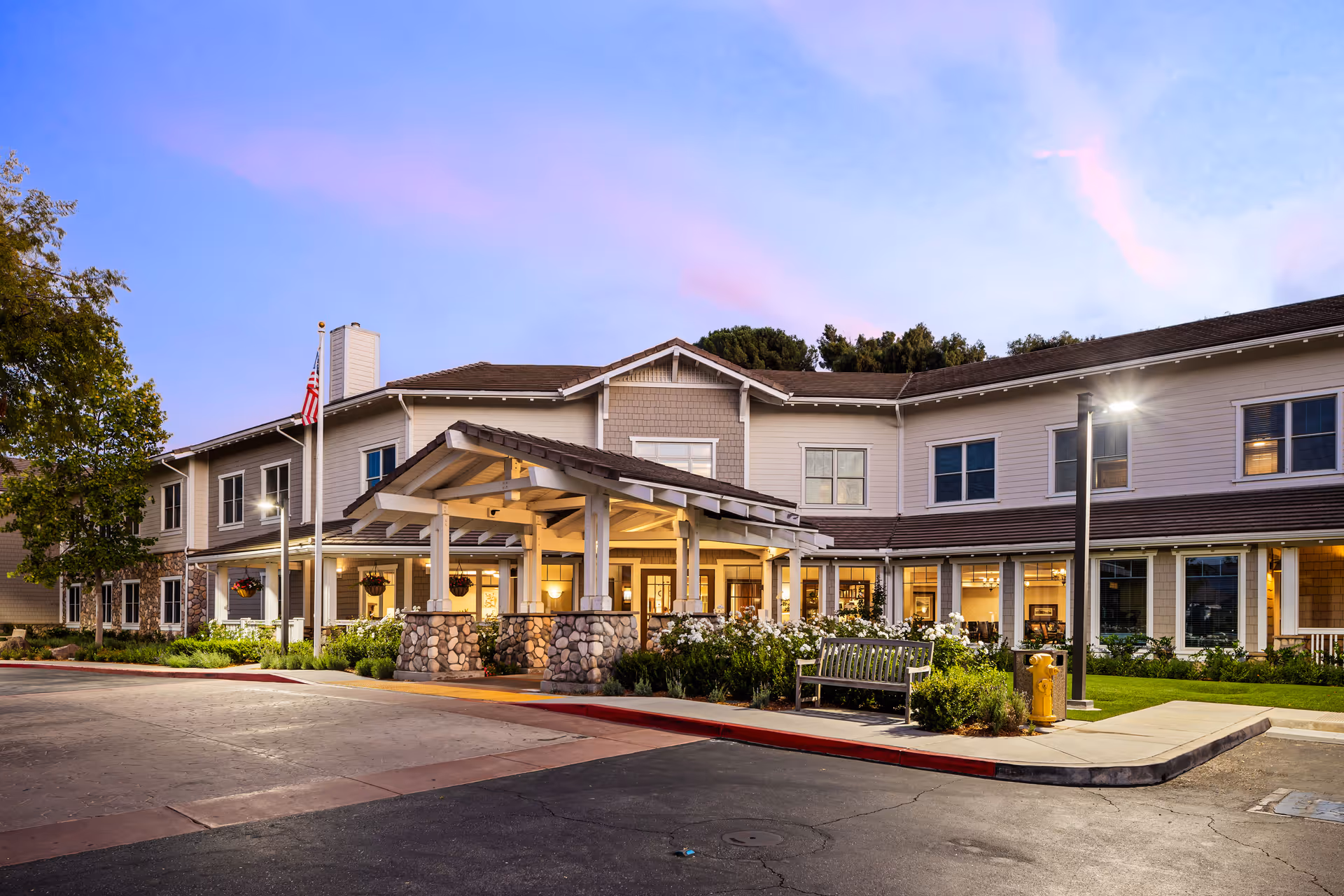 Exterior view of a two-story senior living facility building at dusk with a covered entrance, stone pillars, an American flag, well-maintained landscaping, and a bench near the sidewalk.