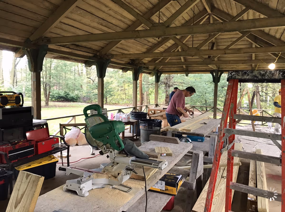 Covered outdoor workspace with wooden beams and roof, featuring a man working on wooden planks at a table. Various woodworking tools and equipment, including a green miter saw, red ladder, toolboxes, and wooden boards are visible. The background shows a green wooded area.