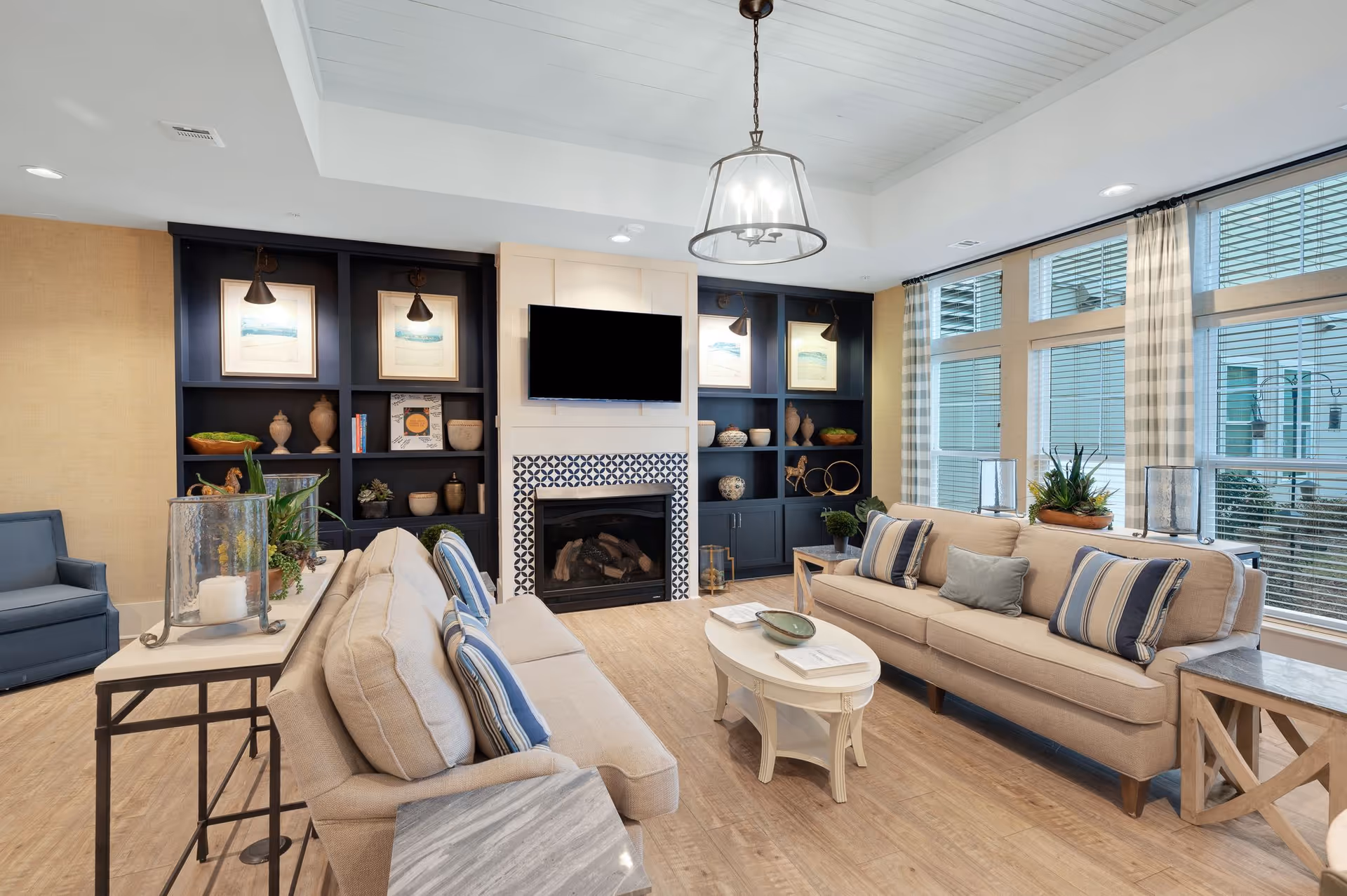 A bright and spacious living room with two beige sofas adorned with striped and solid pillows, a white oval coffee table, and a blue armchair. The room features large windows with checkered curtains, built-in dark blue shelves decorated with vases and framed artwork, a fireplace with a patterned tile surround, and a mounted flat-screen TV above it. The floor is light wood, and a modern pendant light hangs from the white paneled ceiling.