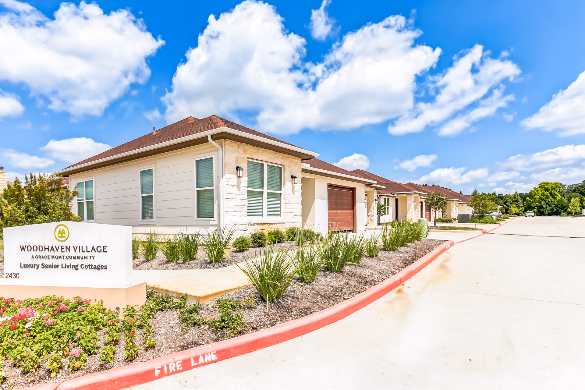 Exterior view of Woodhaven Village, a luxury senior living community featuring single-story cottages with beige siding and stone accents, surrounded by landscaped greenery and a clear blue sky with scattered clouds.