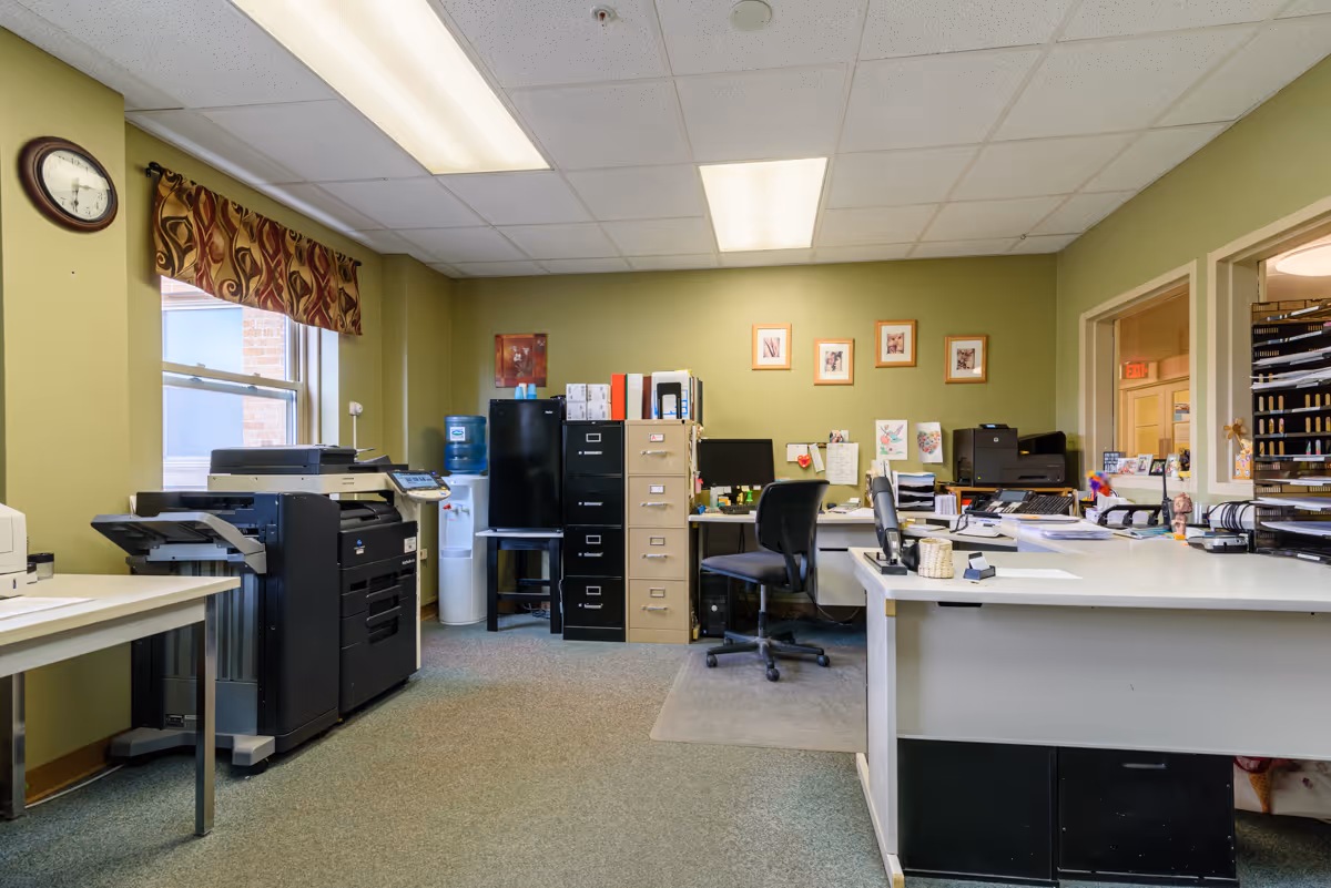 Office workspace with desks, filing cabinets, printers, and chairs in a green-walled room.