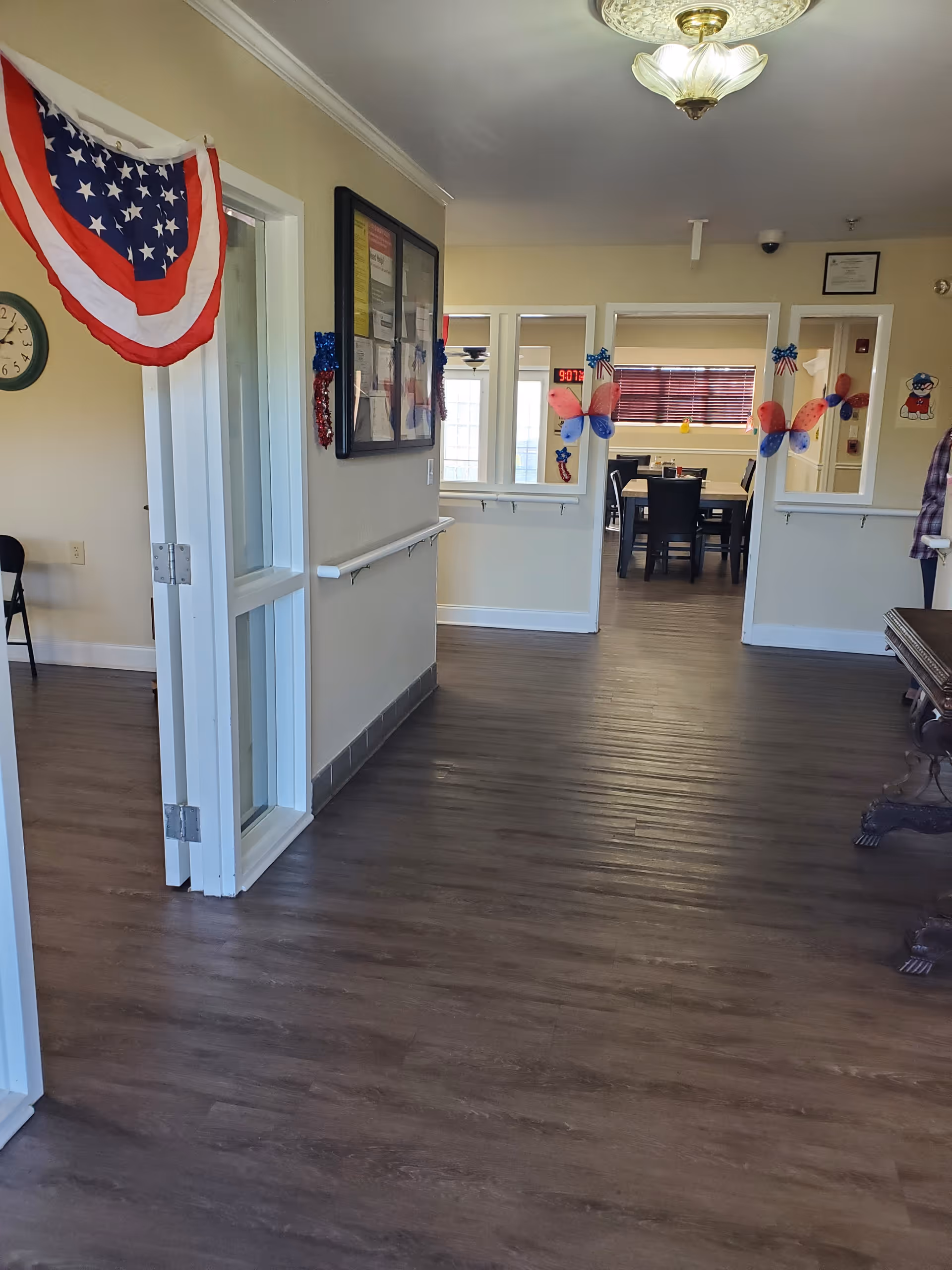 Interior hallway of a senior living facility decorated with patriotic red, white, and blue decorations including a bunting and butterfly-shaped ornaments. The hallway has wood flooring, light-colored walls, and a ceiling light fixture. There are windows and doorways leading to a dining area with tables and chairs visible in the background.