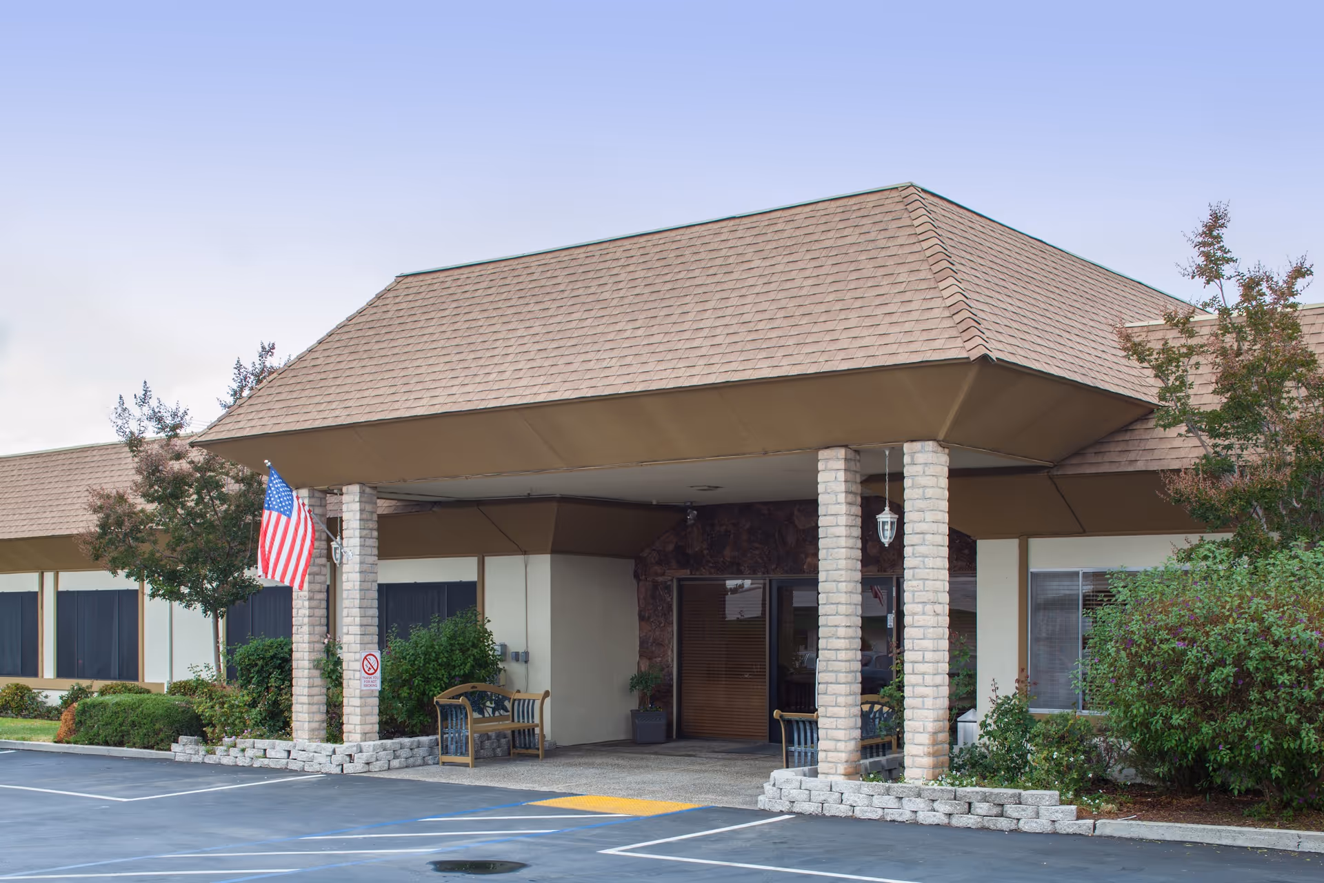 Entrance of a single-story building with a covered porch supported by brick columns. There are benches on either side of the entrance, an American flag hanging from one of the columns, and some bushes and trees around the building. The parking lot is visible in the foreground.