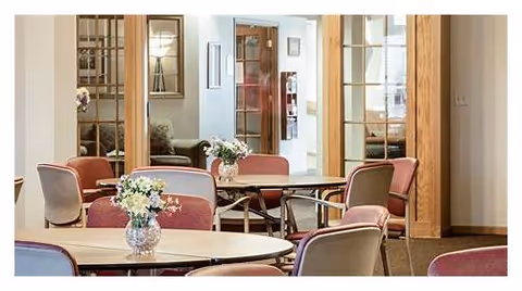 Interior view of a common area with round tables and chairs arranged around them, each table decorated with a small vase of flowers. The background shows glass doors and windows with a glimpse into another room with seating.