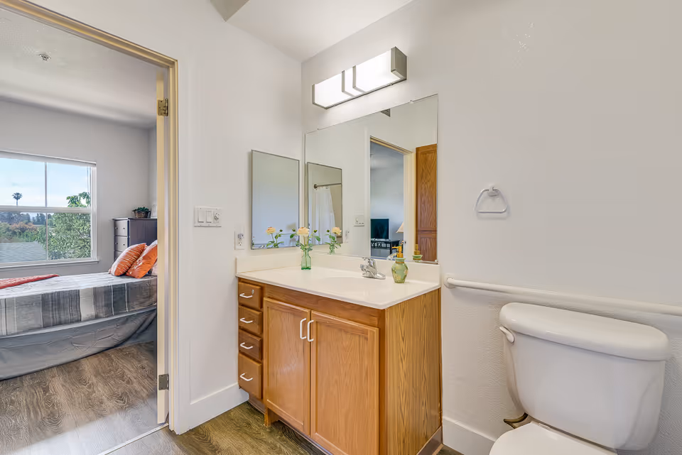 Bathroom with a wooden vanity, sink and mirror, toilet, and an open door showing an adjacent bedroom.
