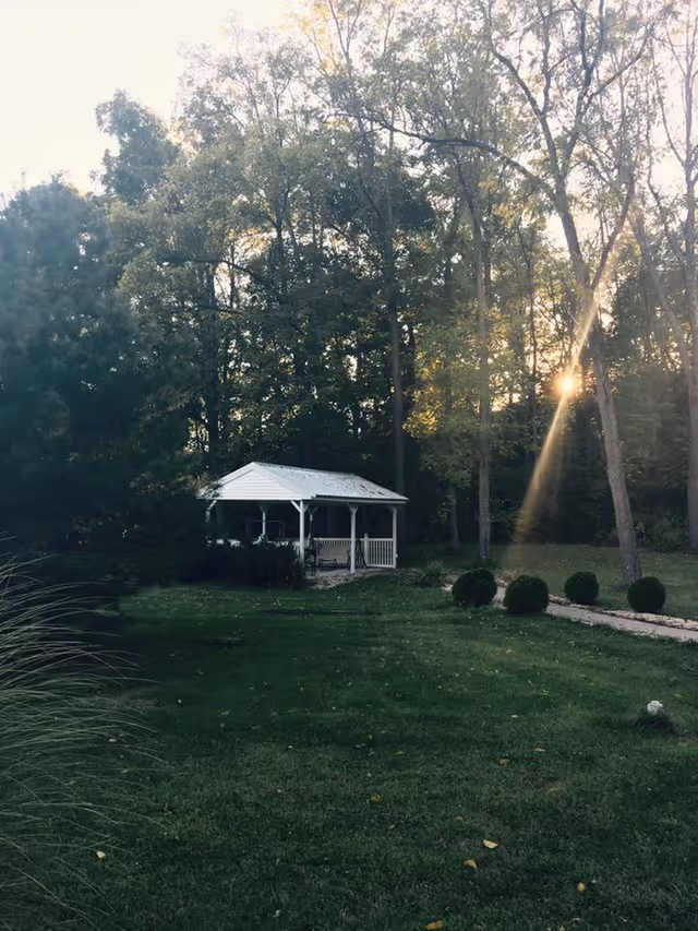 A peaceful outdoor scene featuring a white gazebo surrounded by green grass and trees with sunlight filtering through the branches.