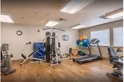 A small fitness room with exercise equipment including a treadmill, stationary bike, multi-function weight machine, and various exercise balls. The room has wooden flooring, white walls, a clock, and windows with blinds allowing natural light.