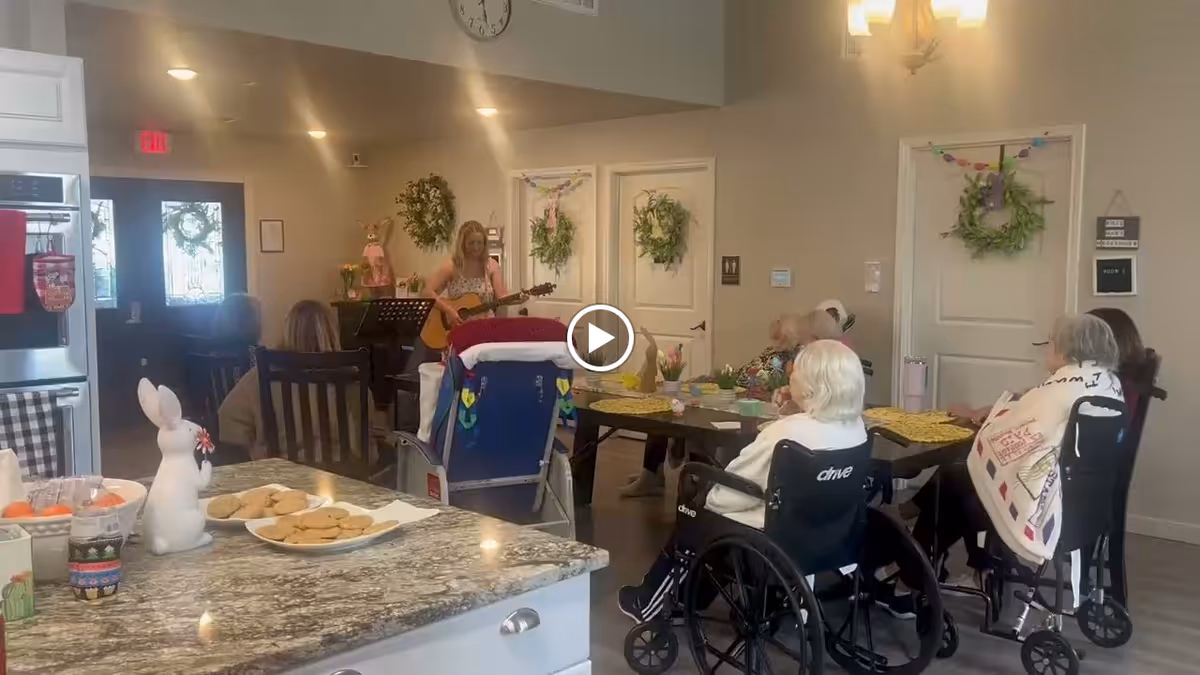 A group of elderly residents, some in wheelchairs, seated around a table in a common area of an assisted living facility. A woman is playing a guitar and singing in front of them. The room is decorated with wreaths on the doors and has a cozy, welcoming atmosphere. In the foreground, a kitchen counter with a plate of cookies and a white ceramic bunny is visible.