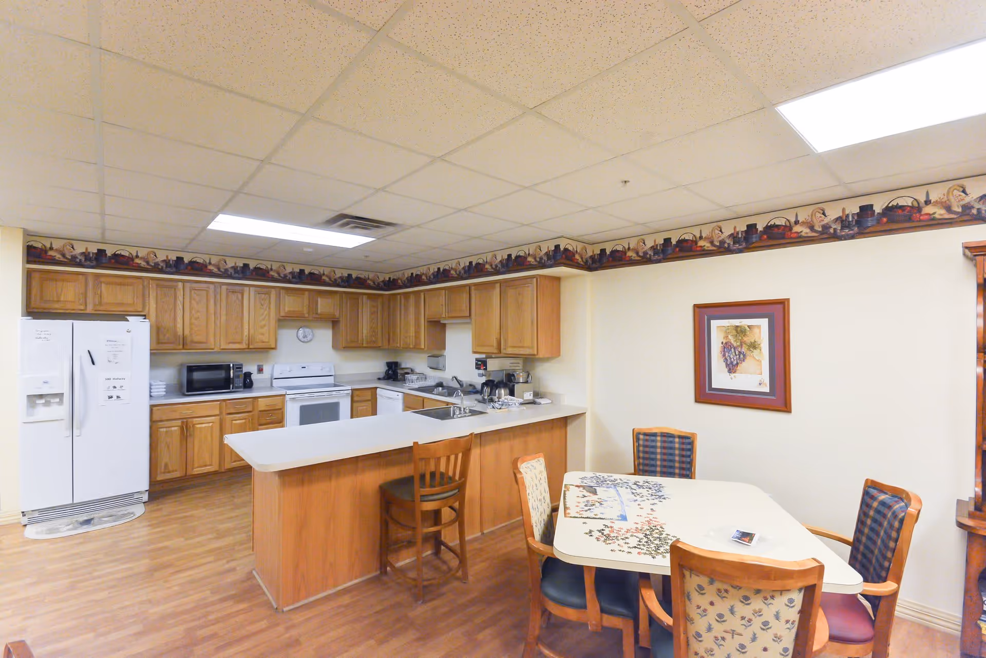 A kitchen and dining area in an assisted living facility featuring wooden cabinets, a white refrigerator, microwave, stove, and dishwasher. There is a countertop with a wooden chair and a dining table with four chairs, two of which have patterned upholstery. The walls are light-colored with a framed picture of grapes and a decorative border near the ceiling.