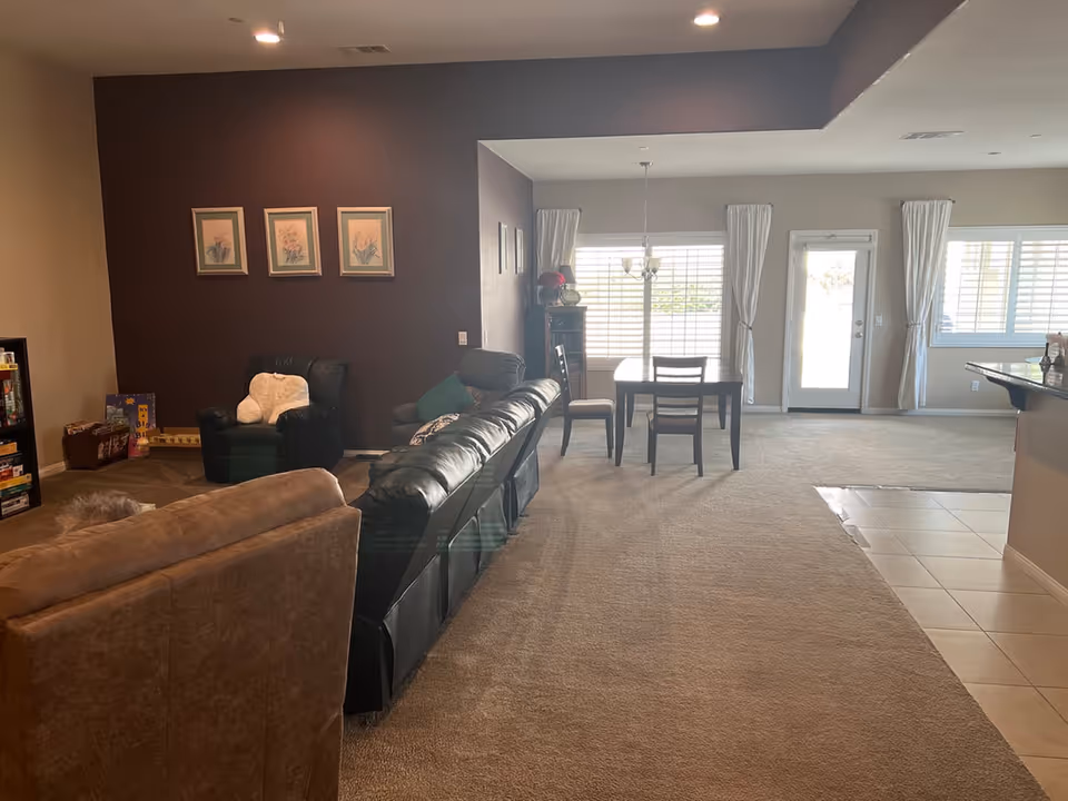 Interior view of a living and dining area in an assisted living facility. The living room features a brown couch, a black leather recliner, and a black armchair with a white pillow. There are three framed floral pictures on a dark brown accent wall. The dining area has a wooden table with four chairs, positioned near large windows with white curtains and a glass door leading outside. The floor is carpeted except for a tiled section near the kitchen counter on the right.